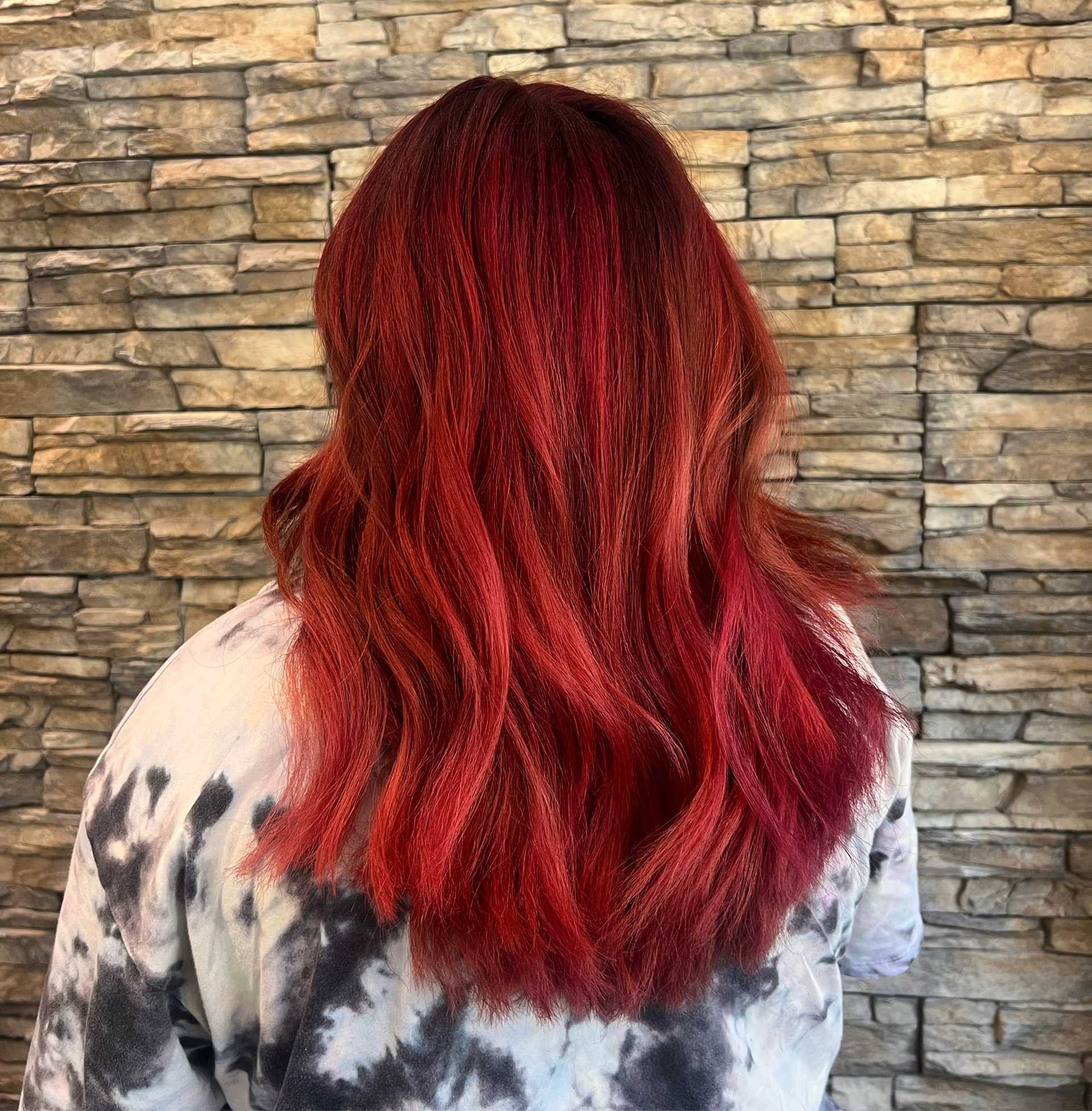 Red hair with dark roots styled in loose waves against a textured stone wall backdrop.